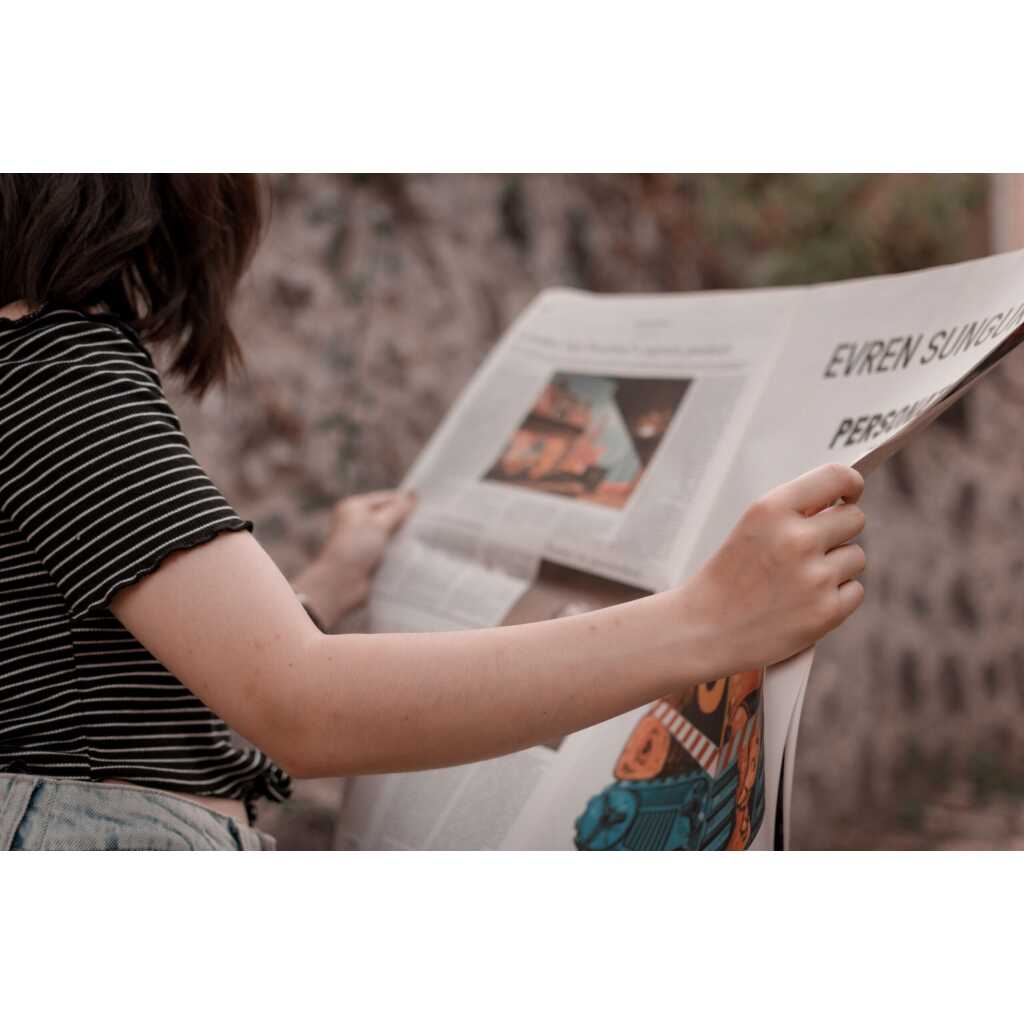 A woman holding a newspaper while sitting outside, enjoying a leisurely read.